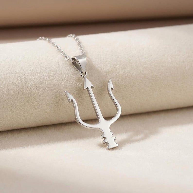 Puede incluir: Un collar con colgante plateado con un dise&ntilde;o de tridente. El tridente tiene una punta central en forma de flecha y dos puntas laterales curvas. La cadena del collar tambi&eacute;n es plateada. El colgante probablemente est&aacute; hecho de acero inoxidable o metal similar.