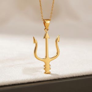 Puede incluir: Un collar con colgante dorado con un dise&ntilde;o de tridente. El tridente tiene tres p&uacute;as curvas y una punta central en forma de flecha. La cadena del collar tambi&eacute;n es dorada y cuelga de un peque&ntilde;o lazo en la parte superior del colgante.