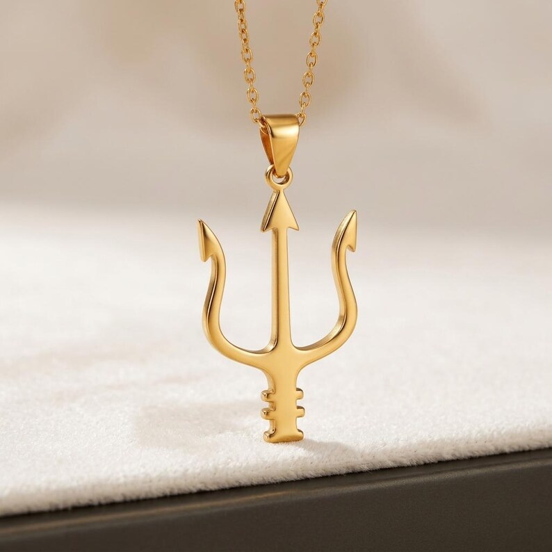 Puede incluir: Un collar con colgante dorado con un dise&ntilde;o de tridente. El tridente tiene tres p&uacute;as curvas y una punta central en forma de flecha. La cadena del collar tambi&eacute;n es dorada y cuelga de un peque&ntilde;o lazo en la parte superior del colgante.