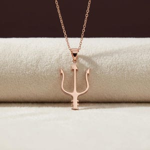 Puede incluir: Collar con colgante de tridente en oro rosa. El colgante presenta un dise&ntilde;o de tridente de tres puntas, suspendido de una delicada cadena. El collar se muestra sobre un fondo neutro, destacando el color y el dise&ntilde;o de la joya.