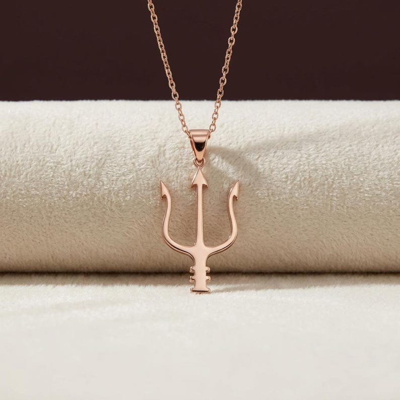 Puede incluir: Collar con colgante de tridente en oro rosa. El colgante presenta un dise&ntilde;o de tridente de tres puntas, suspendido de una delicada cadena. El collar se muestra sobre un fondo neutro, destacando el color y el dise&ntilde;o de la joya.