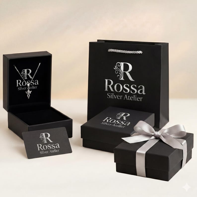 Puede incluir: Un conjunto de cajas de regalo negras y una bolsa de la compra con el logotipo plateado "Rossa Silver Atelier". Una caja est&aacute; abierta, revelando un collar de plata. Una caja de regalo est&aacute; atada con una cinta plateada.