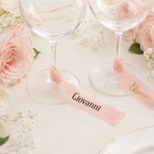Può includere: Due calici in vetro trasparente con steli avvolti da nastri rosa pallido dai bordi sfilacciati. Un nastro presenta il nome Giovanni stampato in nero, mentre l'altro mostra il nome Eleonora stampato in oro. I bicchieri sono circondati da rose.
