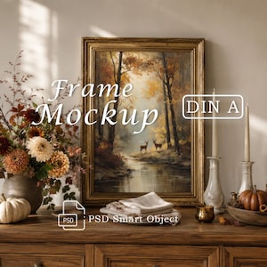 May include: A gold-toned rectangular picture frame containing an art print of a forest scene with two deer near a stream. The image features the text Frame Mockup, DIN A, and PSD Smart Object. The frame is displayed on a wooden surface.