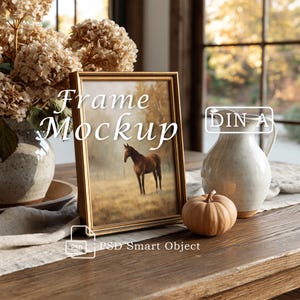 May include: A gold-toned picture frame mockup featuring a horse in a field, with the text Frame Mockup, DIN A, and PSD Smart Object. The frame is displayed next to a ceramic pitcher, a small pumpkin, and a vase of dried flowers.