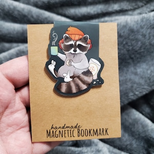 May include: A handmade magnetic bookmark featuring a cartoon raccoon wearing an orange beanie and holding a mug of coffee and a cookie. The raccoon is sitting with two small white rabbits. The bookmark is on a brown card with the text "handmade MAGNETIC BOOKMARK".