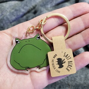 May include: A clear acrylic keychain featuring a cartoon green frog with a grumpy expression. The keychain is attached to a gold metal ring and a brown paper tag with the text "SALT BIRCH . MMM . SALT BIRCH . COM" and a black silhouette of a bat.