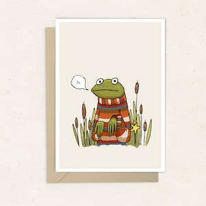 May include: Greeting card featuring a whimsical illustration of a frog wearing a striped sweater and holding a star-shaped wand. The frog is in a marsh setting, with cattails and the word "hi" in a speech bubble.