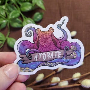 May include: A sticker featuring a stylized octopus with purple and blue tentacles, a pink and orange body, and the word "HYDRATE" on a wooden plank. The sticker has a white border and is held by a hand.