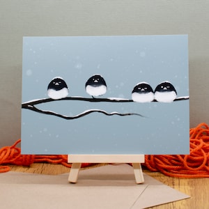 May include: A print of four black and white birds perched on a snow-covered branch against a light blue background. The artwork is displayed on a small wooden easel, with orange yarn and a brown envelope in the foreground.