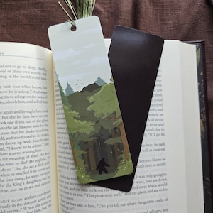 May include: A bookmark featuring a green and brown illustration of a forest with a large bear walking through the trees. The bookmark is printed on a white background and has a black magnetic strip on the back.