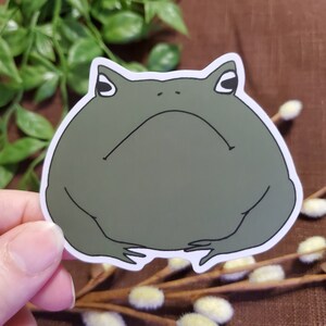 May include: A green frog sticker with a grumpy expression. The frog is sitting on its hind legs with its front legs folded in front of it.