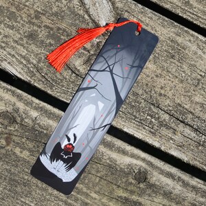 May include: Black bookmark with a red tassel. The bookmark features a stylized illustration of a creature with large eyes and wings in a dark forest setting.
