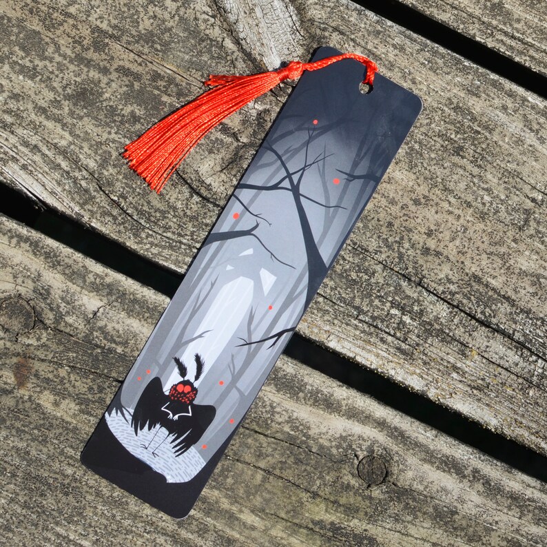 May include: Black bookmark with a red tassel. The bookmark features a stylized illustration of a creature with large eyes and wings in a dark forest setting.