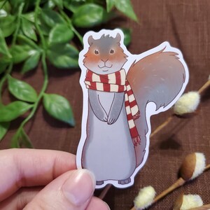 May include: A gray squirrel wearing a red and white striped scarf. The squirrel is standing on its hind legs with its tail curled behind it.
