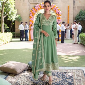 May include: A sage green long-sleeved A-line kurta dress featuring a V-neckline with intricate gold embroidery on the bodice, cuffs, and hem. The outfit includes matching slim-fit trousers and a coordinating dupatta draped over one shoulder.