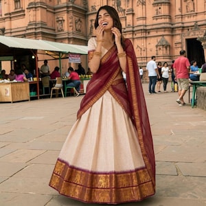 May include: A woman wearing a traditional Indian outfit, a cream-colored skirt with a gold and burgundy border, and a matching burgundy shawl. The outfit is worn with gold bangles. The background features a building and people.