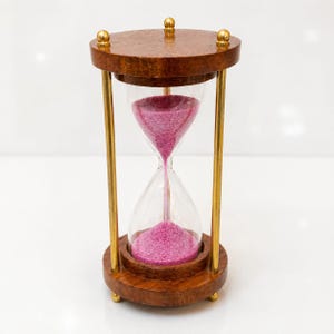 May include: An hourglass with a wooden frame and brass-coloured supports. The hourglass contains pink sand, and the top and bottom are made of dark brown wood. The glass is clear, and the sand is flowing from the top to the bottom.