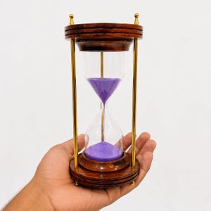 May include: An hourglass with a wooden frame and brass detailing. The hourglass contains purple sand. The wooden frame is dark brown, and the brass accents are gold. The hourglass is held in a hand.