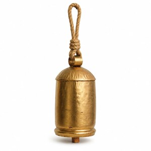 Puede incluir: Una campana de metal de color dorado con cuerpo cilíndrico y parte superior redondeada. La campana presenta una superficie texturizada y martillada, y está suspendida por un lazo de cuerda de yute gruesa de color natural.
