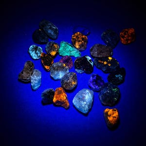 Puede incluir: Una colección de piedras minerales en bruto e irregulares que muestran colores fluorescentes vibrantes bajo luz ultravioleta. Las piedras exhiben tonos brillantes de naranja neón, turquesa, morado intenso y azul eléctrico.