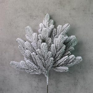 May include: A single artificial evergreen branch with a frosted, snow-covered appearance. The branch features multiple needles and a dark brown stem. The background is a solid gray, creating a contrast with the white and green of the branch.
