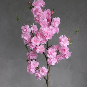 May include: A branch of artificial cherry blossoms in full bloom. The flowers are a vibrant pink, with multiple layers of petals. The branches are brown, and the background is a solid gray.
