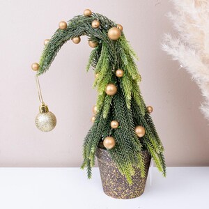 May include: A tabletop artificial Christmas tree with a curved top, featuring green pine needles and matte gold spherical ornaments. A single gold glittery ornament hangs from the curved tip. The tree is set in a speckled dark brown pot.