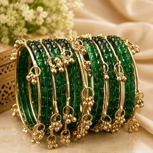 May include: A set of emerald green glass bangles featuring a faceted texture and gold-toned metal accents. Each bangle is adorned with small, dangling gold-toned charms that create a jingling effect. The bracelets are arranged in a stack.