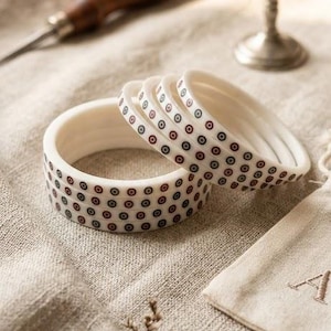 May include: Ivory-colored bangles with a repeating pattern of red, navy, and black dots. The bracelets are stacked on a neutral-toned linen surface. A small canvas bag with the letter "A" embroidered on it is also present.