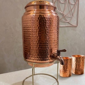 May include: A hammered copper water dispenser with a matching lid and a metal spigot, resting on a gold-toned wire stand. Two matching hammered copper tumblers are positioned next to the dispenser, which features a polished finish.