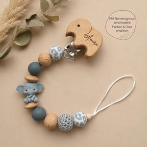 Può includere: Un portaciuccio con una clip in legno a forma di elefante incisa con il nome Amira, collegata a una serie di perline nei toni del grigio, bianco e legno naturale. Le perline includono silicone liscio, uncinetto testurizzato e motivi leopardati.