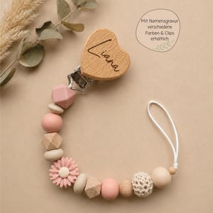 May include: A pacifier clip featuring a heart-shaped wooden piece engraved with the name Liana, attached to a metal clip. The chain consists of various beads in shades of pink, beige, and cream, including geometric shapes and a flower-shaped bead.