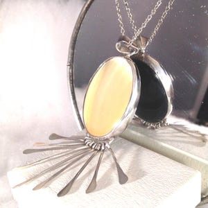 Puede incluir: Un collar con colgante de plata con una piedra ovalada de color crema engastada en un marco de plata. El colgante tiene elementos plateados radiantes que se extienden desde la base. El collar se muestra frente a un espejo, que refleja un colgante negro similar.