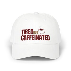 May include: A white baseball cap with the words "TIRED BUT CAFFEINATED" in maroon lettering. A small illustration of a coffee cup with steam rising from it is next to the word "BUT". The cap has a curved brim and a button on top.