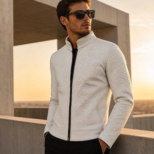 May include: A white textured jacket with a high collar and a black zipper. The jacket is paired with black pants and sunglasses. The background features a concrete structure and a sunset.