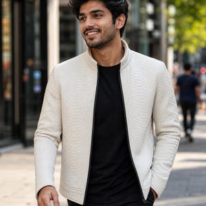May include: A cream-coloured jacket with a textured snakeskin pattern, stand-up collar, and black zip. The jacket is worn over a black t-shirt. The person is walking on a city pavement.