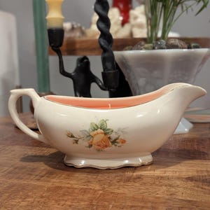 Puede incluir: Una salsera de cerámica color crema reutilizada como portavelas, con un diseño floral central de rosas amarillas y hojas verdes. El interior está relleno de cera naranja claro con una mecha, y presenta bordes dorados en la base y el borde.