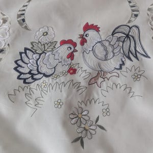 May include: A white fabric cloth featuring detailed embroidery of a rooster and a hen facing each other. The birds are depicted in shades of white, gray, and black with red combs. The design includes embroidered flowers, grass, and cutwork edges.