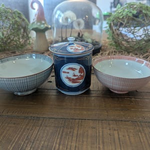 May include: A set of three ceramic pieces including a dark blue lidded tea cup with orange landscape illustrations, a light blue checkered bowl, and a light pink checkered bowl. Each piece features a smooth finish and a rounded, functional shape.