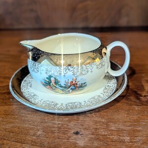 May include: A cream-colored porcelain creamer and saucer set featuring a gold metallic rim and intricate gold filigree patterns. The creamer displays a colorful illustration of two figures in a garden setting, complemented by a matching saucer.