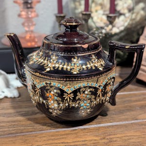 May include: A dark brown ceramic teapot featuring a glossy finish, a matching lid with a knob, and a curved handle. The body is decorated with intricate gold, turquoise, and orange floral patterns and beaded accents along the rim and base.