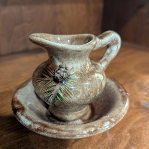 May include: A small brown glazed ceramic creamer pitcher with a matching saucer. The pitcher features a hand-painted design of a pinecone with green pine needles on its side. The glaze has a mottled, variegated brown finish throughout.
