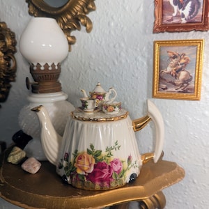 May include: A white ceramic teapot featuring a fluted body with a colorful floral pattern of yellow, pink, and red roses. The lid is topped with a miniature teapot, a tiny teacup on a saucer, and a small sugar bowl, all accented with gold trim.