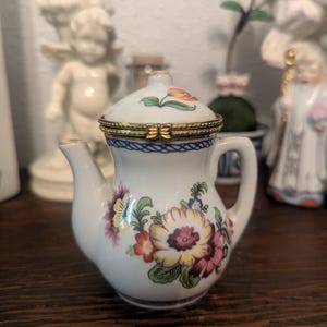 Könnte beinhalten: Eine kleine Teekannen-Schmuckdose aus weißem Porzellan mit einem bunten Blumenmuster aus gelben, rosa und grünen Blüten. Der Deckel ist mit einem goldfarbenen Metallverschluss in Schmetterlingsform versehen und zeigt ein kleines Tulpenmotiv.