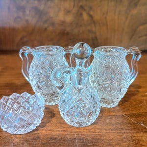 May include: A collection of four clear pressed glass items including two small handled pitchers with a starburst pattern, a small round bowl with a diamond-cut texture, and a small cruet bottle with a matching stopper and a diamond-cut base.