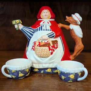 Może przedstawiać: Ceramiczny czajniczek w kształcie Czerwonego Kapturka w czerwonym kapturze i pelerynie, trzymający koszyk z czerwono-białą szachownicą. Brązowa figurka wilka tworzy uchwyt. W zestawie znajdują się dwie małe, pasujące filiżanki w żółte kwiatowe wzory.