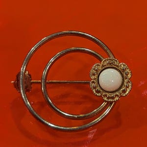 Op de afbeelding: Een ronde broche bestaande uit twee concentrische ringen van zilverkleurig metaal. Aan de rechterkant van de ringen is een kleine, goudkleurige, bloemvormige zetting bevestigd met een gladde, ronde, witte cabochonsteen in het midden.
