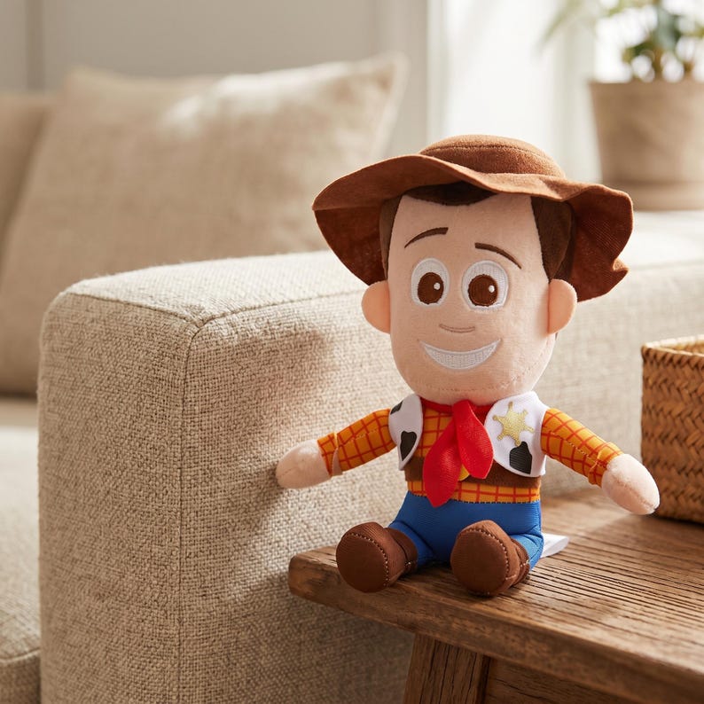 May include: A plush toy character featuring a brown cowboy hat, a yellow and orange plaid shirt, a red neckerchief, a white vest with black cow-print spots and a gold star badge, and blue pants. The figure has a soft fabric face with brown eyes.