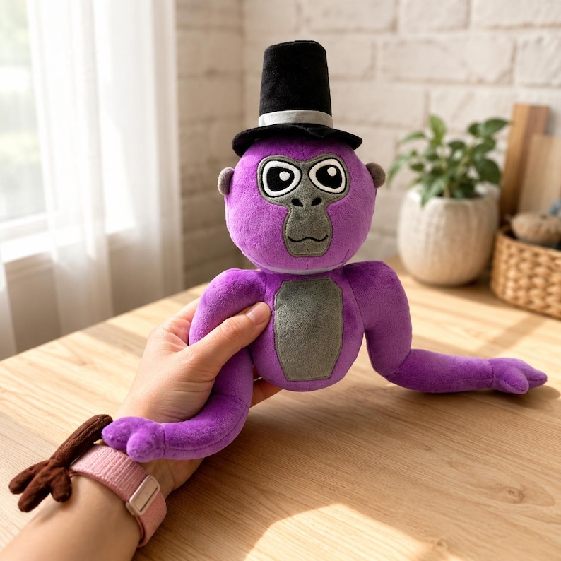 May include: A plush purple gorilla toy featuring a grey face and chest patch, wearing a black top hat with a silver band. The stuffed animal has long arms and large eyes with white highlights, crafted from soft fabric with visible stitching details.